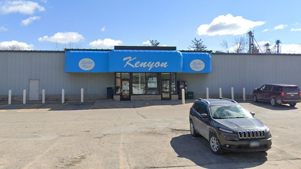 The storefront from Red Wing Avenue: gray steel building, blue awning with the Kenyon script, parking in front